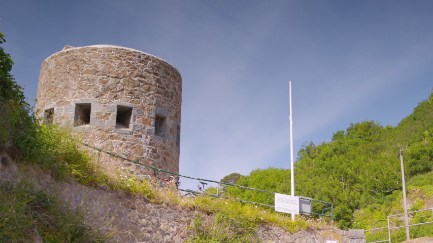 A daylight medium shot of the exterior facade of a round cylindrical tower made of stone and surrounded with wild plants.