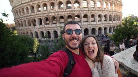 Happy young tourist couple making selfie in front of colosseum in rome, italy. Beautiful smiling people in honeymoon traveling in Italy. Unidentified tourists walking behind
