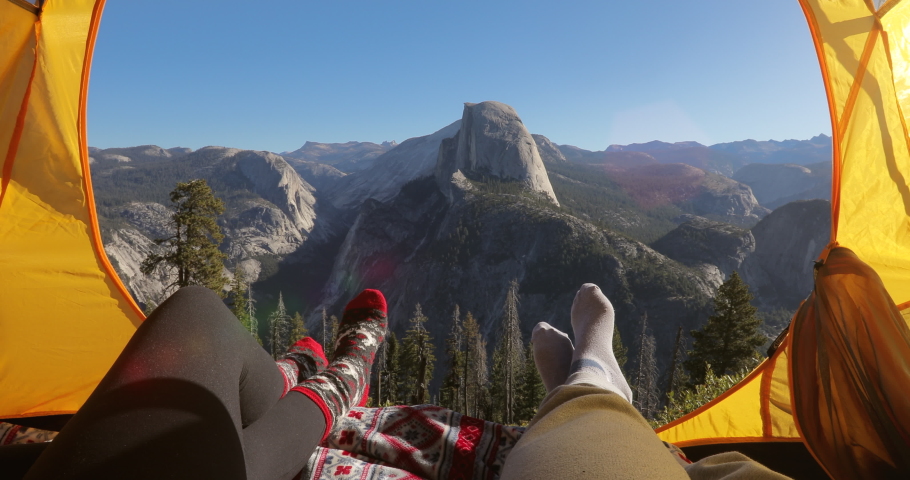 Two pairs of legs in the opening of the tent, against the backdrop of the mountain landscape of Sierra Nevada and the cliff of Half Dome.The sun illuminates the tent.Outside landscape looks inspiring.