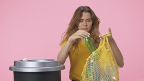 A good-looking young woman starts to sort garbage then puts her string bag to the litter isolated over pink background