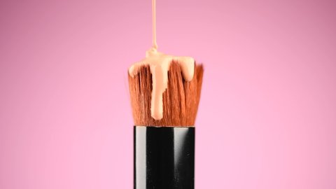 Make-up liquid foundation pouring on makeup brush, closeup. Foundation beauty facial cosmetics, Kabuki tool for perfect make up. Dripping bb cream or concealer, over pink background. 4K slow motion