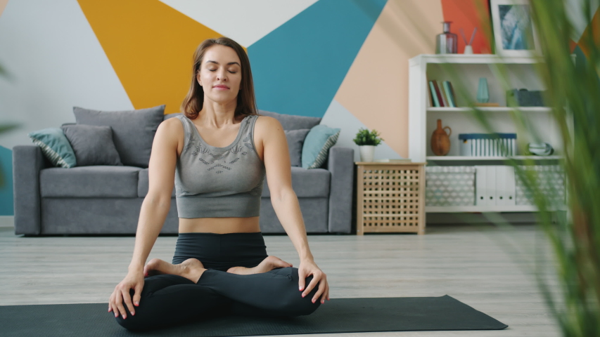 Relaxed young woman in sportswear is meditating in lotus position sitting on yoga mat with closed eyes at home enjoying meditation with hands in namaste. Royalty-Free Stock Footage #1043351545