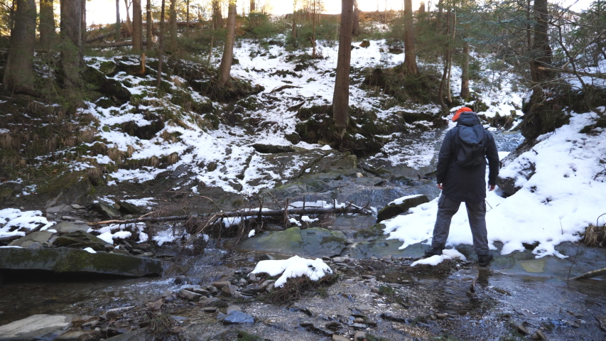 Young backpacker walks over stones on mountain river at pine forest. Unrecognizable hiker with backpack goes over rocks on small stream during travel. Concept of winter vacation or holiday. Slow mo
