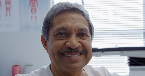 Portrait close up of a senior mixed race man looking straight to camera and smiling in a hospital examination room, mustached, window background