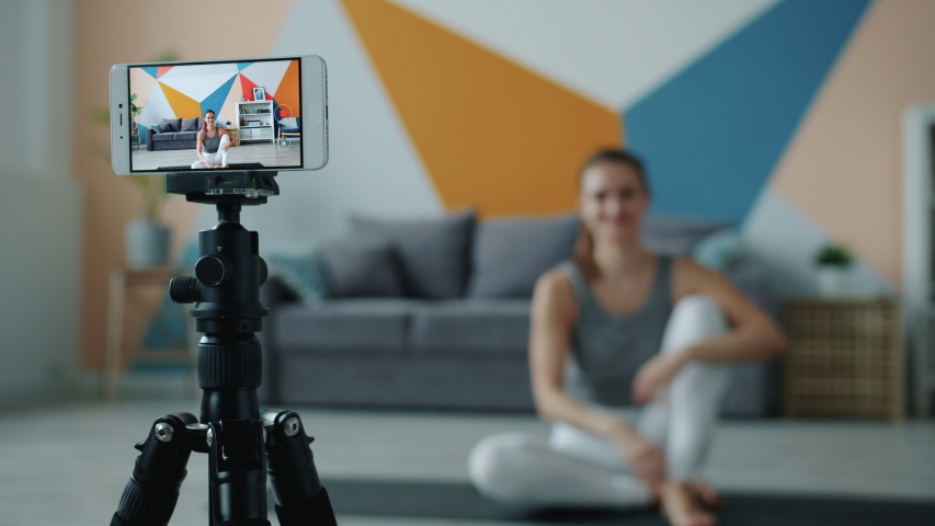 Female blogger is recording video with smartphone camera doing hatha yoga at home, focus is on mobile screen on tripod. Modern technology and vlogging concept. Royalty-Free Stock Footage #1043356903