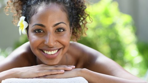 Closeup face of young african american woman in a wellness center ready for massage therapy. Portrait of beautiful girl lying down on massage table at spa resort. Smiling girl looking at camera.