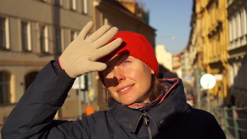 Young woman in red hat squinting from the bright winter sun on European old street on winter day handheld shot