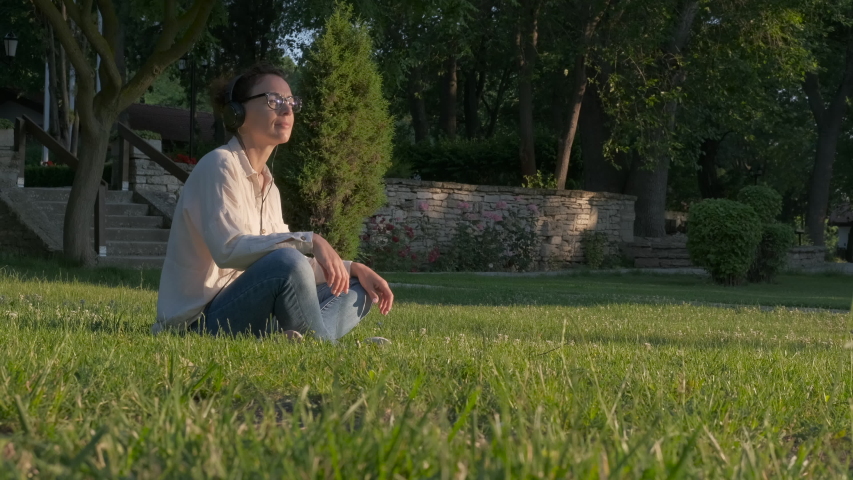 In headphones in the park on the grass. A woman is enjoying music on a green lawn.