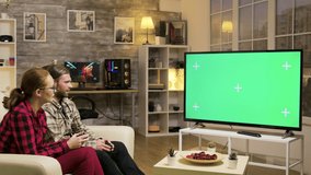 Relaxed couple sitting on sofa playing video games using wireless joysticks on tv with green screen.