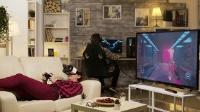 Woman lying on sofa playing video games using vr headset in living room. Boyfriend playing on computer.