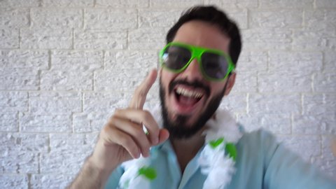Happy man comes up from below dancing and singing to the camera. Carnival reveler is wearing green sunglasses, white and green flower necklace, light blue casual shirt. Close up, face.
