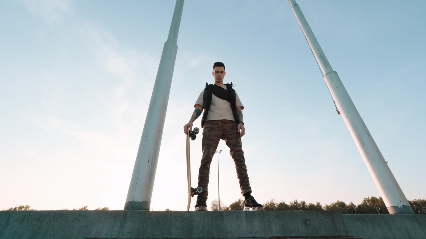 Young Guy Stands On The Street With Skateboard In His Hands. Guy Stands Between Two Electric Poles. Sunlight Illuminates The Guy.