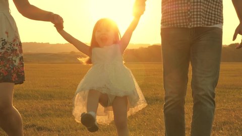 child plays with dad and mom on field in sunset light. Walking with small kid in nature. little daughter jumping holding hands of dad and mom in park on background of sun. Family concept.