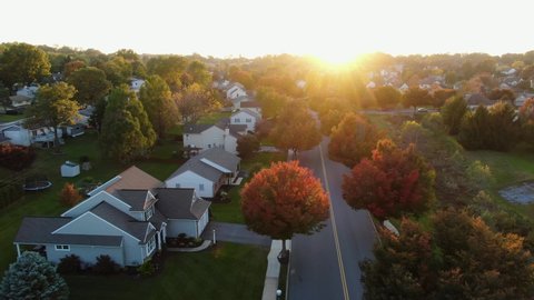 Sunset starburst highlights autumn neighborhood scene with homes and colorful trees, cars and trucks on street at intersection, aerial drone shot