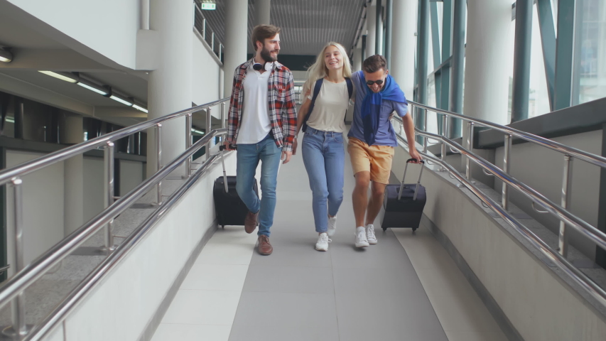 Three funny friends with baggage walking along airport building before or after check-in, having talk, smiling, hugging. Happy hipster tourists walking to train ready to start their trip together