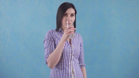 young woman with bewilderment drinks water from a glass standing on a blue background