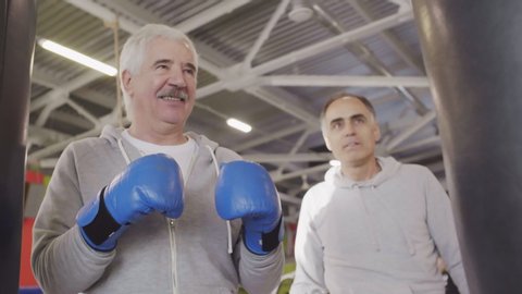 Close up of retired Caucasian sporty man wearing boxing gloves exercising with boxing bag while his middle-aged teammate standing nearby and watching. Then they making air punch