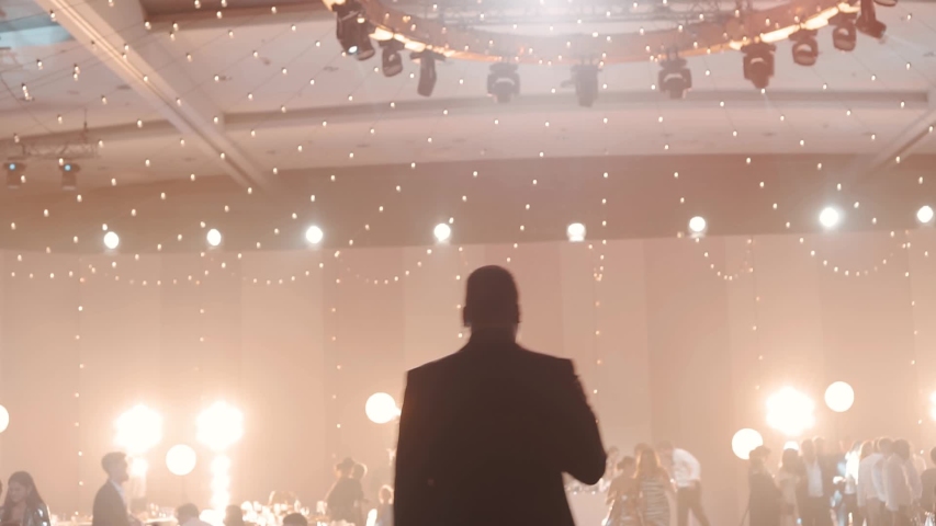 Silhouette of a singer of a young man on a background of listeners, lights, spotlights, rays of light. The man speaks from the stage and thanks the audience. Taken from the back.