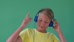 Closeup view of handsome white young kid listening to music standing near green wall background. Kid using blue headphones. Real time 4k video footage.