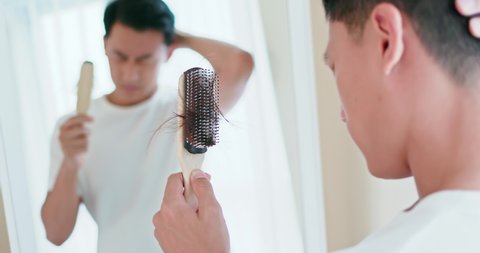Asian man worry about his receding hairline and look in mirror with many hair on hairbrush