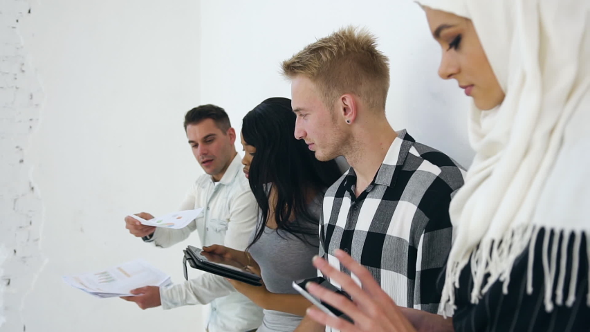 Likable contented 30s mixed race office colleagues standing close to the white wall and working with financial documents with charts