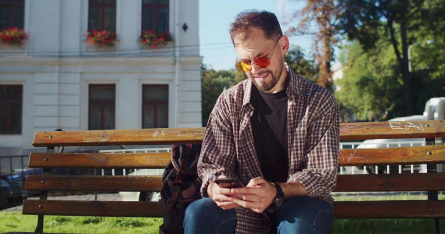 Happy young man learning exciting news using smartphone clenching fist like enjoying success or victory sitting on bech in the downtown.