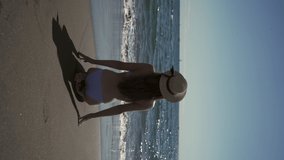 A beautiful girl in a swimsuit and a straw hat stands on the ocean on a sandy beach and admires the view in sunny summer weather. Rest and travel during working holidays. Vertical video.