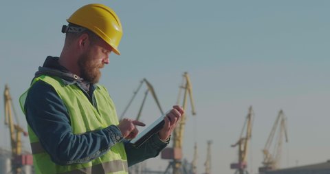 Port Worker With A Beard In A Yellow Helmet Stands With A Tablet PC In The Seaport  Against The Background Of Cranes. The Foreman Inspects The Industrial Harbor. Cinema 4K Video