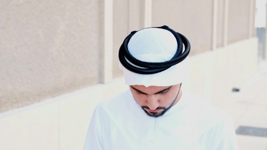 Arab man looks up to the camera smiling. Happy Arabic Emirati national smiles as he looks in front wearing Kandura. Middle Eastern guy on dish dasha UAE menswear | Shutterstock HD Video #1043471827