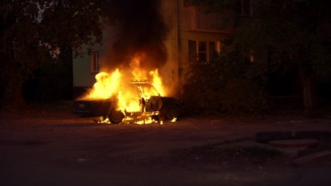 Burning car close to a house in a sleeping area of the city. Theme of riots criminal arson of a car near house. Automobile engulfed in flames of fire. Trees close to burning sedan.
