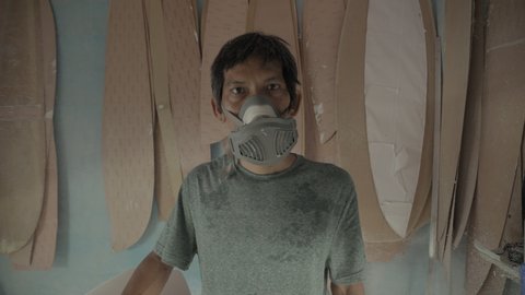 Zoom out of professional surfboard shaper wearing protective mask standing in workshop and looking at camera