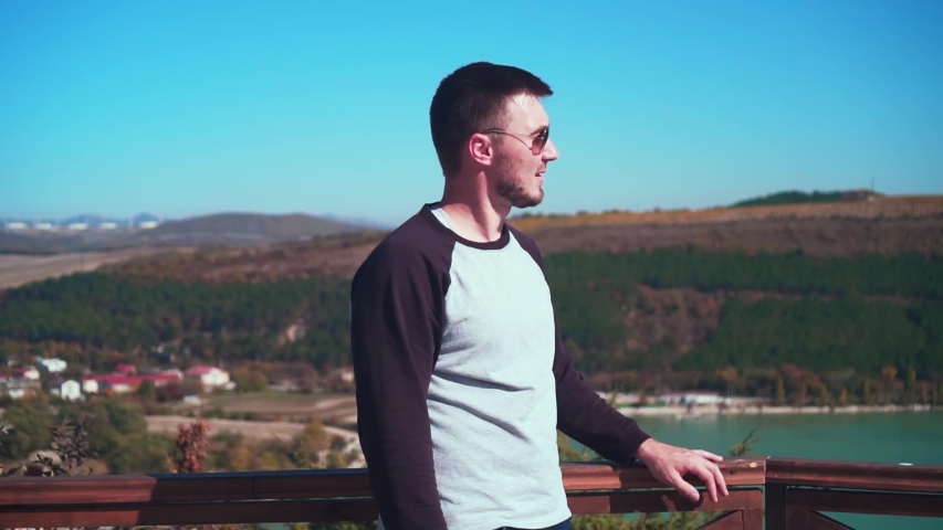 A young man is standing, looking from the observation deck and bouncing up, raising his hands and showing super signs and peace from happiness, joy of victory, success and achievement.