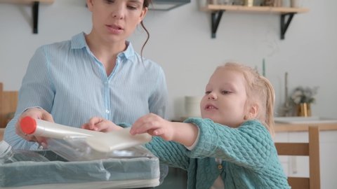 A little girl, about 5 years old, puts plastic in a special basket. Mom teaches daughter to sort waste for recycling in a bright kitchen. Garbage sorting concept.
