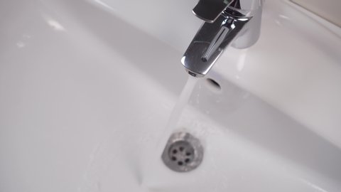 A man’s hand closes a faucet in a white bathroom sink and tap water stops pouring. White clean sink. 