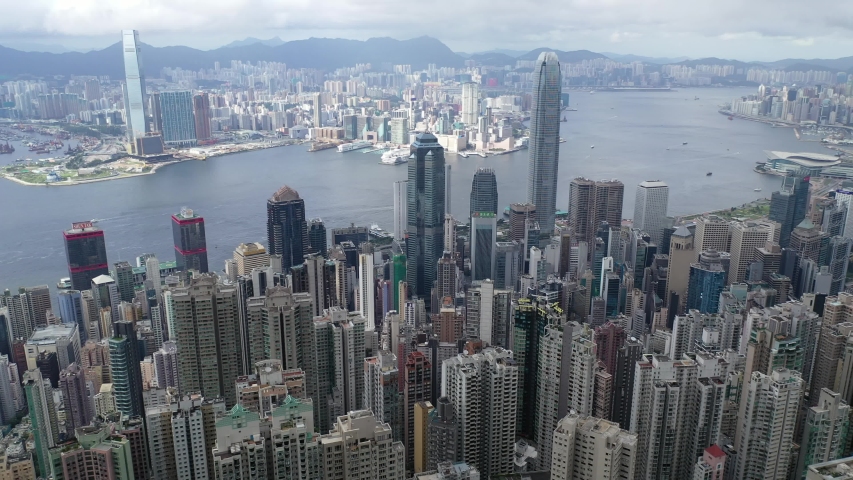 Aerial panorama of Hong Kong viewed from top of Victoria Peak with skyscrapers on HongKong Island & in Kowloon Downtown by Victoria Harbour & residential towers on Mid-Levels hillside under cloudy sky