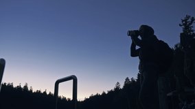 Silhouette traveler photographing scenic view in forest, river. Wood pier. One woman shooting nice dark magic night look. Girl take photo video on camera. Photographer walk with backpack. Outdoor.