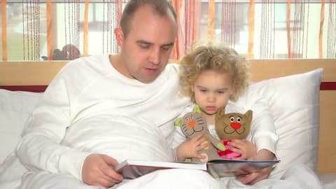 Playful dad reading book and playing with his young daughter girl sitting under wrap in bed. Static shot.