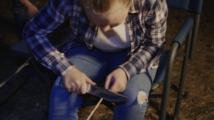 Teenage girl whittling twig with knife in forest camp at night. Beautiful teenage girl in checkered shirt holding knife and sharpening wooden stick in dark camping
