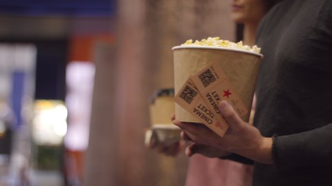 Tracking of young woman with two cups of coffee to-go walking alongside unrecognizable man holding popcorn and movie tickets. Couple ready to watch film at cinema