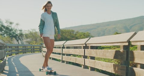 Young attractive blonde skater girl riding her longboard skateboard on a coastal boardwalk, smiling skateboarder cruising with style