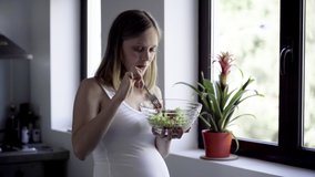 Cheerful pregnant woman eating fresh salad. Future mother standing near window with salad bowl. Pregnancy and healthy nutrition concept