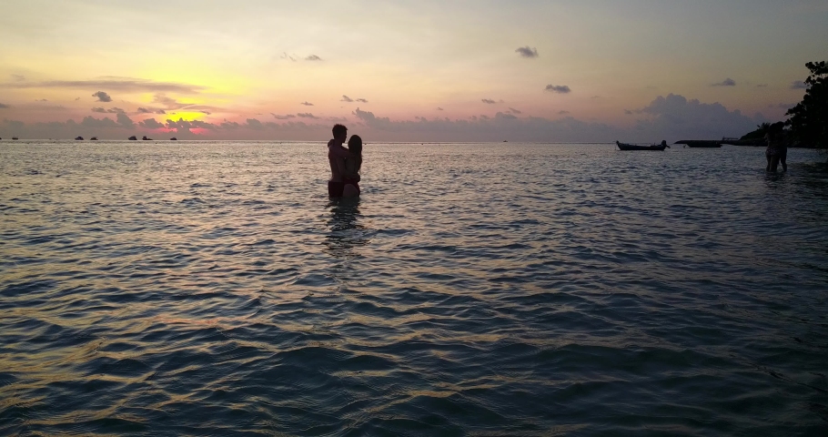 Tourist Romantic Couple Give Sweet Kiss To Each Other And Enjoy the Touristic Island Beach in Greece With Wonderful Orange Sunset At the Background, Golden Hour - Wide Shot