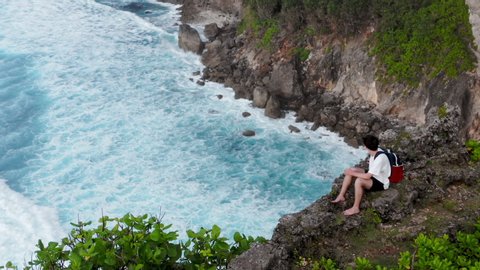 Aerial view of man sitting on a green cliff. Man sitting and looking at horizon. Boy comes to edge of crag and sits on rock. Lonely guy sitting over cliff. Indian Ocean, Uluwatu, Bali, Indonesia.