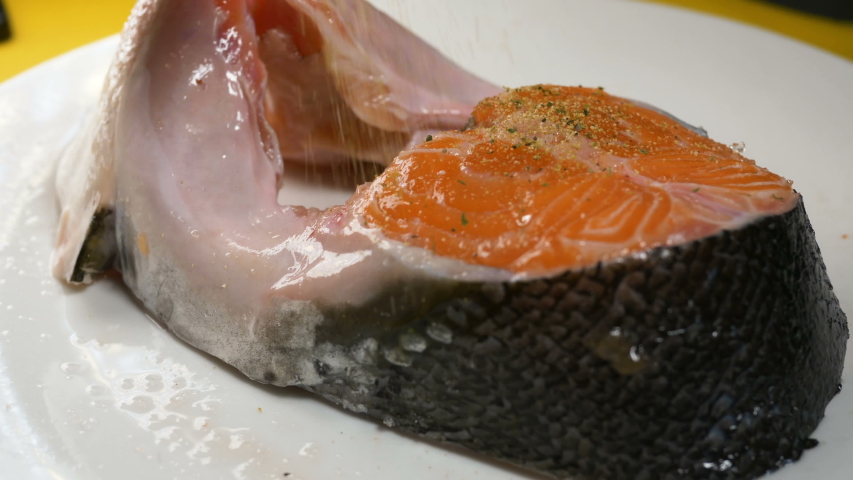 person sprinkles large red fish piece with various spices on white plate in restaurant kitchen extreme close view
