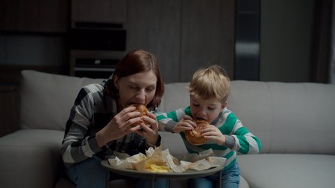 Family eating fat burgers sitting on sofa. Brunette mother and preschool blonde son eating meal at home. 