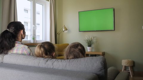 Family Sitting Together Sofa In Their Living Room Watching TV Green Screen. Rear View Of Family With Children Sitting On Sofa In Living Room Day Watching Green Mock-up Screen TV Together.