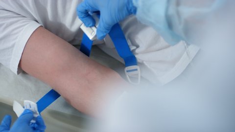 Top view of nurse in disposable gloves applying tourniquet to arm of male patient, disinfecting his skin with alcohol wipe, inserting the needle and placing tape over it while giving IV therapy