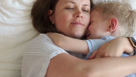 Portrait of tender mom with little son in bed in the morning, they woke up and hugging. Mom hugs her beloved son lying in bed. Slow motion.