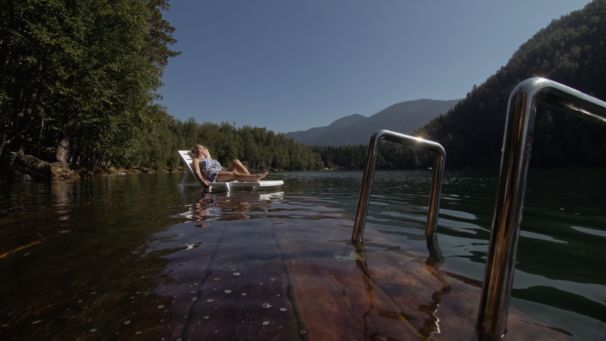 Woman lies on a sunbed in sunglasses and a boho silk shawl. Girl rests on a flood wood underwater pier. The pavement is covered with water in the lake. In the background are mountain and a forest.