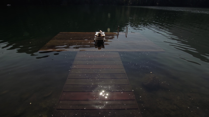 Woman lies on a sunbed in sunglasses and a boho silk shawl. Girl rest on a flood wood underwater pier. The pavement is covered with water in the lake. In the background are a mountain and a forest.